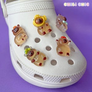 Puede incluir: Un zapato blanco de zueco con varios amuletos de animales coloridos unidos al zapato. Los amuletos están hechos de un material plástico translúcido y presentan diseños de oso, león y otros animales. Los amuletos están unidos al zapato utilizando los agujeros del zapato.