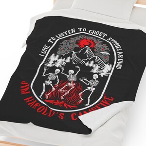 May include: Black and white blanket with a design featuring skeletons dancing around a campfire. The blanket has the text "I LOVE TO LISTEN TO GHOST STORIES AROUND JIM HAROLD'S CAMPFIRE". The design includes mountains, trees, and a red sun.