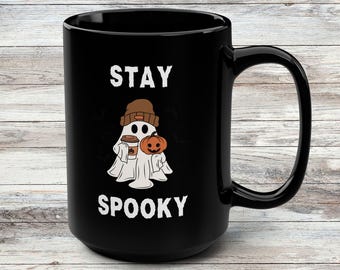 Op de afbeelding: Een zwarte keramische mok met een witte afbeelding van een geest met een bruine muts, die een koffiekopje en een kleine oranje pompoen vasthoudt. De tekst STAY SPOOKY staat in het wit boven en onder de illustratie van de geest gedrukt.