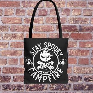 May include: Black tote bag featuring a white graphic with the words "Stay Spooky Campfire", a skull, flames, and the text "Listen to Jim Harold's Campfire". The bag has black handles and is set against a brick wall.