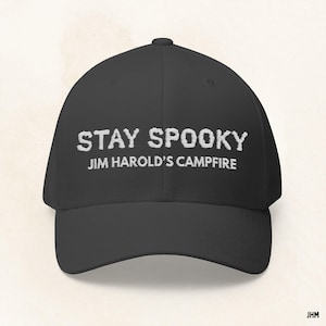 May include: A black baseball cap featuring the embroidered phrase "STAY SPOOKY" above "JIM HAROLD'S CAMPFIRE" in white. The cap has a curved peak and a structured design, ideal for everyday use.