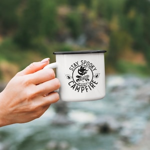 May include: White enamel camping mug with a black rim and handle. The mug has a black and white graphic design that says "Stay Spooky Jim Harold's Campfire".