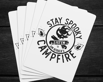 May include: A stack of white playing cards with a black graphic design. The design includes the words "Stay Spooky Campfire" around a central image of a skull and flames. The text "Listen to Jim Harold's" is also included.