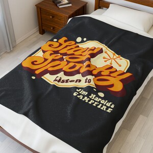 May include: Black blanket with the words "Stay Spooky" in orange and red lettering. The blanket also says "Listen to Jim Harold's Campfire" in white text. The blanket is on a bed with a white pillow and a wooden nightstand.