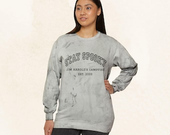 May include: A light gray sweatshirt with the text "STAY SPOOKY" above "JIM HAROLD'S CAMPFIRE EST. 2009" in black. The sweatshirt has a marbled effect and long sleeves. The model is wearing black pants.