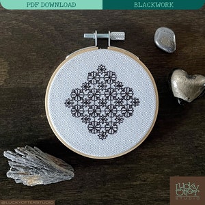 Könnte beinhalten: Schwarzarbeit-Stickmuster eines geometrischen Designs in einem weißen Stickrahmen auf einer dunklen Holzoberfläche. Das Design besteht aus sich wiederholenden schwarzen Blumenformen.