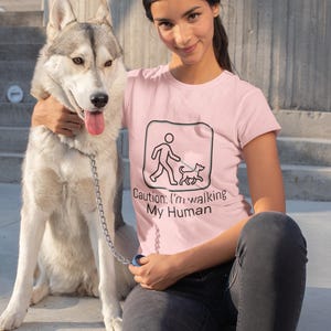 May include: A light pink short-sleeved t-shirt featuring a black graphic of a dog walking a human on a lead inside a rounded square, with the text "Caution: I'm walking My Human" printed in black lettering below the graphic.