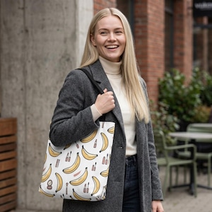 Può includere: Una borsa tote bianca con un motivo ripetuto di banane gialle intervallate da piccole illustrazioni di articoli per il trucco, tra cui rossetti, mascara e specchietti compatti, realizzati in uno stile grafico semplice e pulito.
