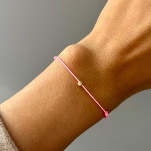 Puede incluir: Una delicada pulsera rosa con una pequeña cuenta dorada. La pulsera se lleva en la muñeca, con un fondo neutro. La pulsera es sencilla y elegante, adecuada para el uso diario.