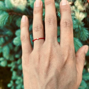 May include: A red string bracelet with a small gold bead on a person's finger.