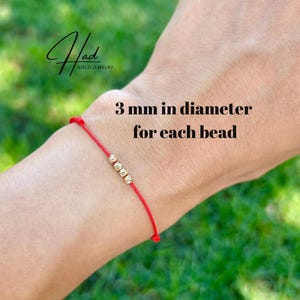 Könnte beinhalten: Ein rotes Schnurarmband mit drei kleinen, runden, texturierten Goldperlen. Der Text auf dem Bild gibt 3 mm in diameter for each bead und Had Gold Jewelry an. Das Armband ist dünn und schlicht, wobei die Goldperlen mittig auf der Schnur sitzen.