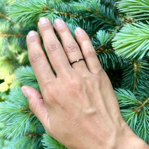 Puede incluir: Un anillo de hilo negro con una pequeña cuenta dorada. El anillo se lleva en un dedo, sobre un fondo de agujas de pino verdes.
