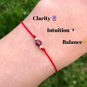 Könnte beinhalten: Rotes Armband mit einer violetten Kristallperle und goldenen Akzenten. Das Armband trägt die Worte "Klarheit", "Intuition" und "Balance".