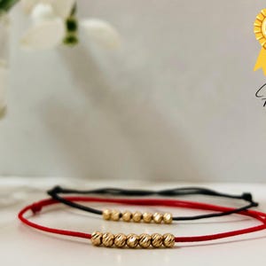Könnte beinhalten: Zwei Armbänder, eines rot und eines schwarz, mit goldenen Perlen. Das rote Armband hat eine Reihe goldener Perlen in der Mitte. Das schwarze Armband liegt auf dem roten Armband. Die Armbänder liegen auf einer weißen Oberfläche. Der Text "14k GOLD" befindet sich auf einem goldenen sternförmigen Aufkleber.