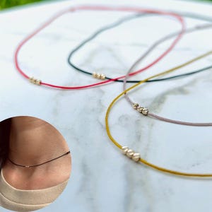 Peut inclure: Plusieurs colliers délicats avec des cordons fins rouges, noirs, gris et jaunes. Chaque collier comporte de petites perles dorées. Un collier est porté autour d'un cou.