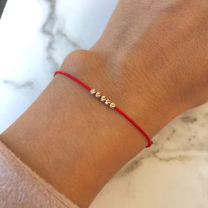Könnte beinhalten: Ein rotes Armband mit kleinen goldenen Perlen. Das Armband ist um das Handgelenk einer Person gebunden.