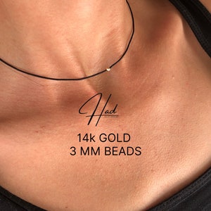 Può includere: Un primo piano di una collana con cordoncino nero con un solo perlino d'oro da 3 mm. Il testo "14k GOLD 3 MM BEADS" è visibile sotto la collana.