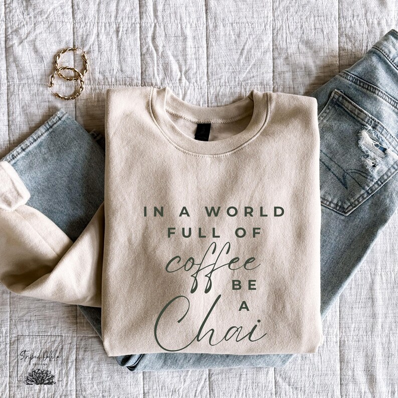 Puede incluir: Una sudadera beige con el texto "In a world full of coffee be a chai" impreso en la parte delantera.