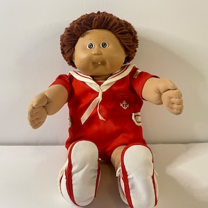 May include: A vintage Cabbage Patch Kid doll with brown yarn hair, wearing a red sailor outfit with a white anchor emblem and white shoes.