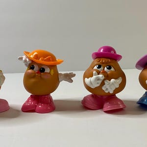May include: Four plastic Mr. Potato Head figures with different hats and facial expressions. The figures are brown with pink, orange, and purple accents.