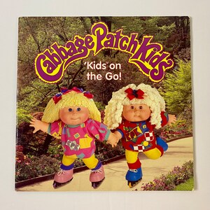 May include: Two Cabbage Patch Kids dolls wearing colorful outfits and roller skates. The dolls are in a green grassy area with flowers and trees. The text on the book cover reads "Cabbage Patch Kids" and "Kids on the Go!"