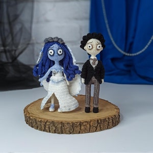May include: Crocheted wedding cake topper featuring a blue-haired bride and a groom in a black suit. The couple is standing on a wooden base.
