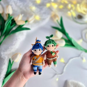 Può includere: Due figure all'uncinetto, una con capelli blu e occhiali che indossa un abito giallo e blu, l'altra con capelli verdi che indossa un abito marrone e verde. Le figure sono tenute in una mano.