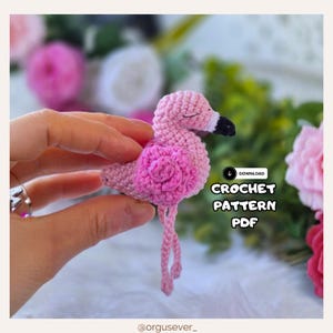 May include: A handmade crochet flamingo in shades of pink, with a darker pink flower detail on its chest. The flamingo has a black and white beak and closed eyes. The text "CROCHET PATTERN PDF" is visible.