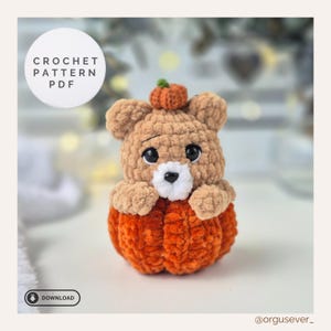 Puede incluir: Un oso de ganchillo sentado dentro de una calabaza naranja. El oso es de color marrón claro con una pequeña calabaza en la cabeza. La imagen incluye el texto "CROCHET PATTERN PDF" y un botón de descarga.