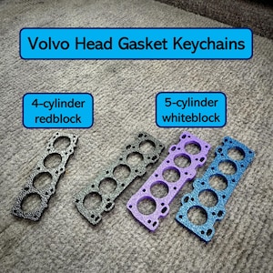 Puede incluir: Cuatro llaveros de junta de culata Volvo. Dos son negros, uno es morado y uno es azul. Los llaveros están etiquetados como "4 cilindros redblock" y "5 cilindros whiteblock".