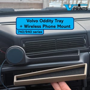 Puede incluir: Salpicadero de coche con una bandeja Volvo Oddity y un soporte para teléfono inalámbrico para la serie 740/940. La bandeja es beige y rectangular, y el soporte para teléfono es un círculo azul oscuro. El salpicadero es gris oscuro.