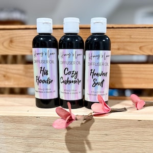 Peut inclure: Trois flacons d'huile de diffuseur noirs avec des bouchons blancs, étiquetés "His Hoodie", "Cozy Cashmere" et "Heaven Sent". Chaque bouteille contient 118 ml. Des fleurs roses sont au premier plan, sur une surface en bois.