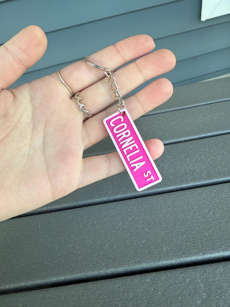 以下が含まれることがあります： 「Cornelia St」の文字が入った、ピンクと白のストリートサインデザインのキーホルダー。キーホルダーはシルバーのキーリングとチェーンに付いています。
