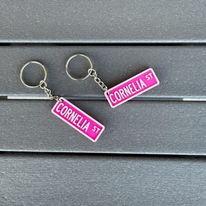 以下が含まれることがあります： 「Cornelia St」と書かれたピンクと白のストリートサインのデザインのキーホルダー2つ。キーホルダーには、シルバーカラーのキーリングとチェーンが付いています。背景は濃いグレーのテクスチャ加工された表面です。