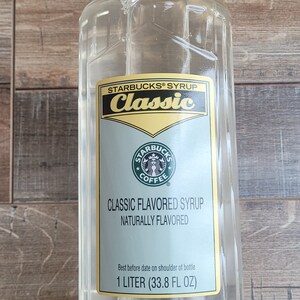 Puede incluir: Una botella de plástico transparente de jarabe con sabor clásico de Starbucks. La botella está etiquetada con el logotipo de Starbucks y el texto "Starbucks Syrup Classic Flavored Syrup Naturally Flavored 1 Litro (33.8 Fl Oz)"