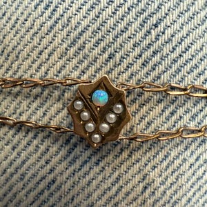 May include: A delicate gold-toned necklace with a central starburst pendant. The pendant features a small, iridescent opal and is surrounded by tiny pearls. The necklace has a double chain design and is set against a blue denim background.
