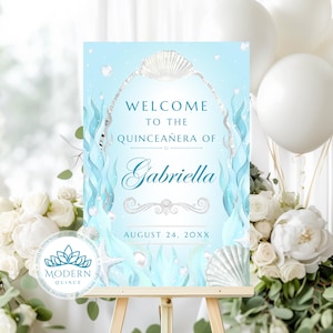 Puede incluir: Un letrero de bienvenida azul claro para una Quinceañera con el nombre Gabriella, fechado el 24 de agosto de 20XX. El diseño incluye un arco de conchas plateado, algas verde azulado, perlas blancas y un adorno decorativo.