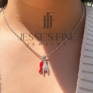 May include: A delicate silver necklace with a small pendant featuring a red chili pepper and a silver charm. The pendant is adorned with small, sparkling stones. The necklace is displayed on a person wearing a white sweater. The text "Jesse's Fine Jewelry" is visible.