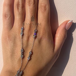 Könnte beinhalten: Eine filigrane silberne Handkette mit tropfenförmigen und runden, klaren Edelsteinen. Das Schmuckstück umfasst eine Ring- und Armbandkette. Der Markenname "Jesse's Fine Jewelry" ist sichtbar.