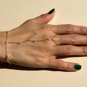 May include: A gold chain bracelet with a small charm at the wrist and four evil eye charms extending to the finger. The bracelet is worn on a hand with green nail polish.