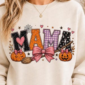 May include: A cream-colored crewneck sweatshirt featuring the word MAMA in large, patterned letters. The design includes a spiderweb, small hearts, stars, two orange pumpkins with pink bows, and a witch's broom tied with a large pink bow.