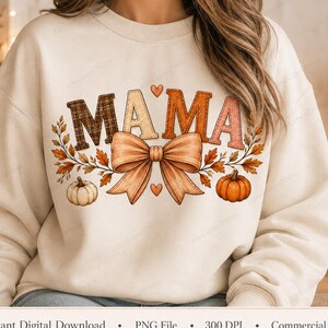 May include: A cream-coloured crewneck sweatshirt featuring the text MAMA in a mix of plaid, solid, and textured patterns. The design includes a large central bow, autumn leaves, and two pumpkins, with small hearts above and below the bow.