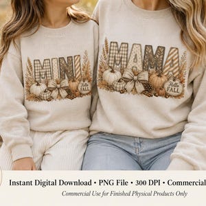 Può includere: Due felpe beige a girocollo con una stampa grafica che riporta le parole MINI e MAMA in un motivo a quadri e righe. Il design include illustrazioni di zucche, spighe di grano, pigne e fiocchi con il testo Hello Fall.
