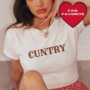 May include: A white short-sleeve t-shirt with the word CUNTRY printed in brown letters across the chest, paired with red and white gingham shorts and red cowboy boots featuring white star and line stitching details.