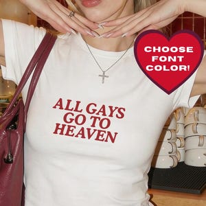 May include: White t-shirt with the text "ALL GAYS GO TO HEAVEN" in red. The shirt is paired with black pants and a burgundy purse. A heart-shaped graphic says "CHOOSE FONT COLOR!"