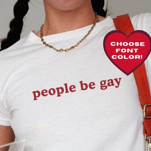 May include: White t-shirt with the phrase "people be gay" in red lettering. A red heart-shaped graphic with the text "CHOOSE FONT COLOR!" is on the right. The person is holding a martini glass with olives.