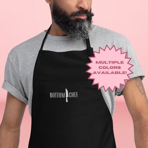 May include: Black apron with the text "Bottom Chef" and a knife graphic. The apron has adjustable straps and a large pocket. A pink starburst sticker says "Multiple Colors Available!"