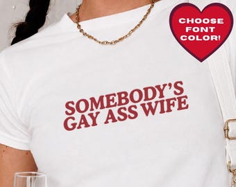 May include: A white short-sleeved t-shirt featuring the red text "SOMEBODY'S GAY ASS WIFE" printed on the chest. The shirt is paired with a gold chain necklace and a white shoulder bag with gold hardware. A red heart graphic displays "CHOOSE FONT COLOR!"