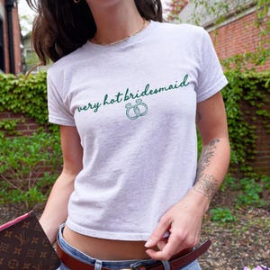 Puede incluir: Camiseta gris claro con la frase "very hot bridesmaid" en verde y dos ilustraciones de anillos verdes. La camiseta tiene mangas cortas y cuello redondo.