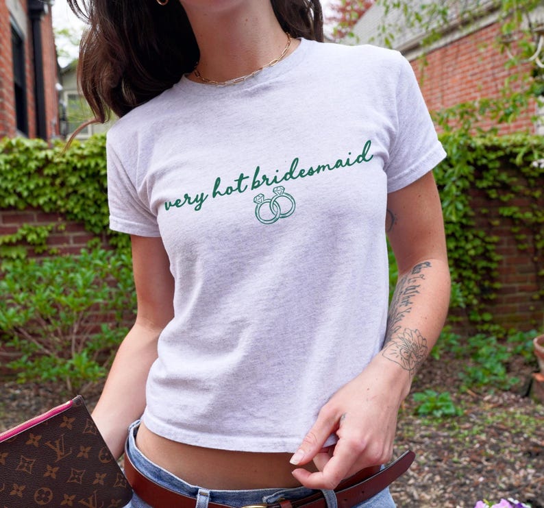 Puede incluir: Camiseta gris claro con la frase "very hot bridesmaid" en verde y dos ilustraciones de anillos verdes. La camiseta tiene mangas cortas y cuello redondo.